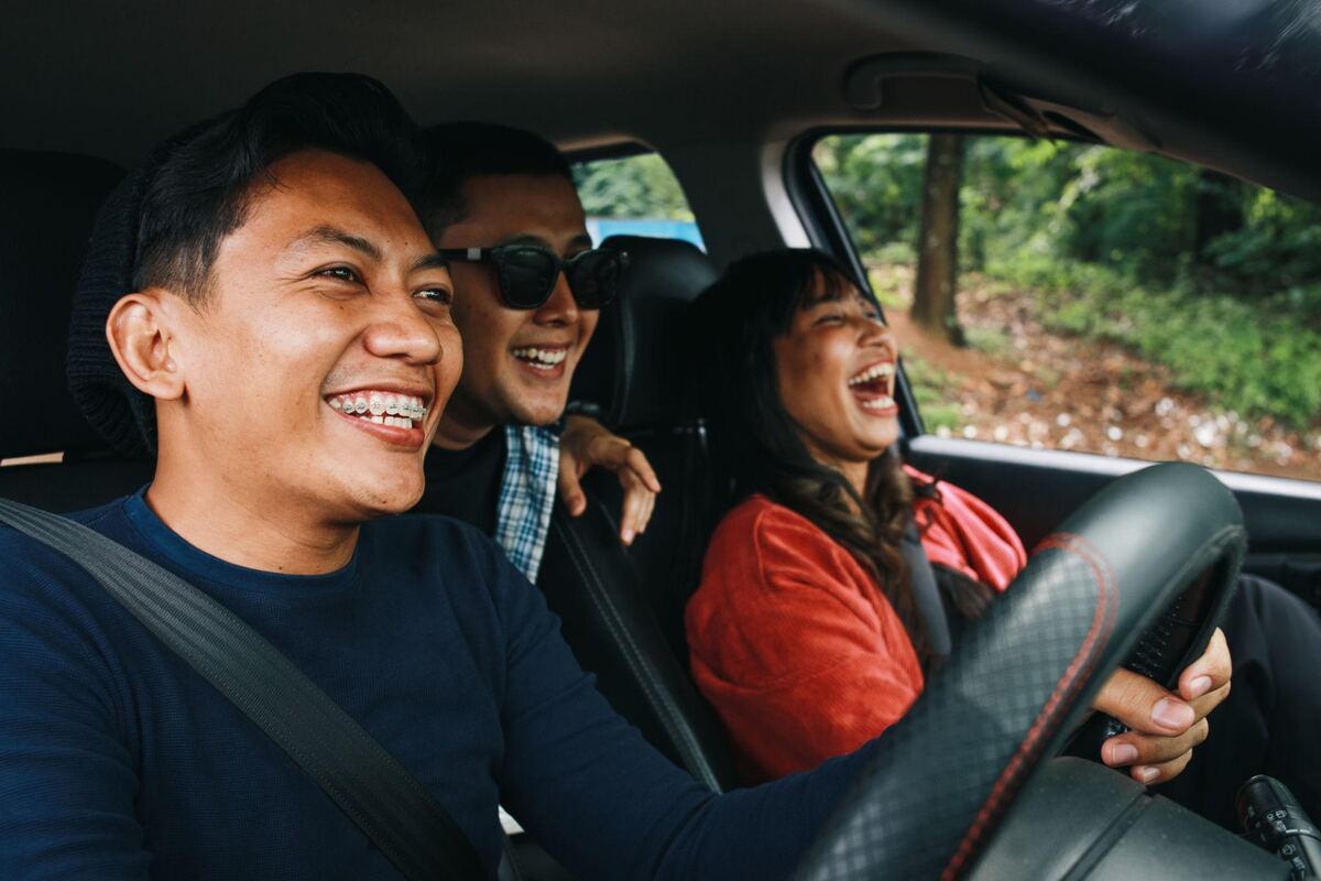 A driver and two friends laughing on a tree-lined road